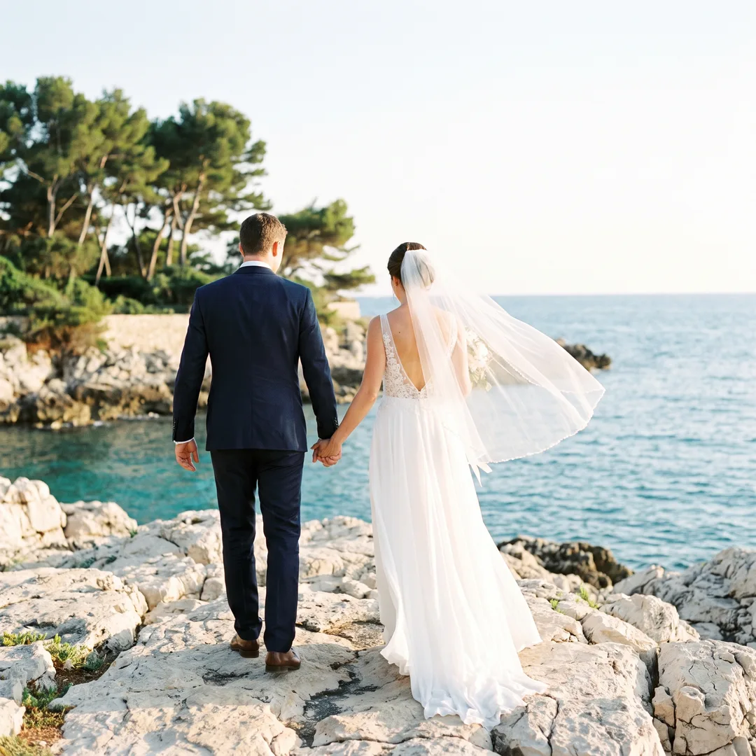 A bride and groom walking on the pale rocks of the Cap d'Antibes coastal path at golden hour, the sea turquoise below