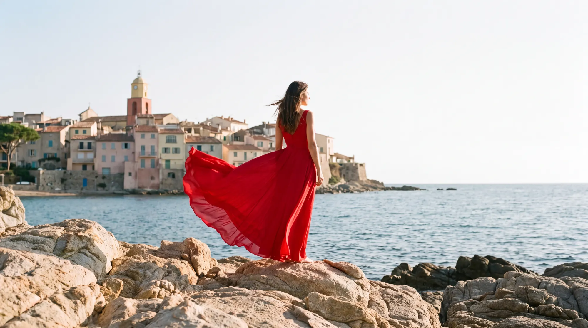 A woman in a long red dress on the coastal rocks near Saint-Tropez, the train lifted by the wind