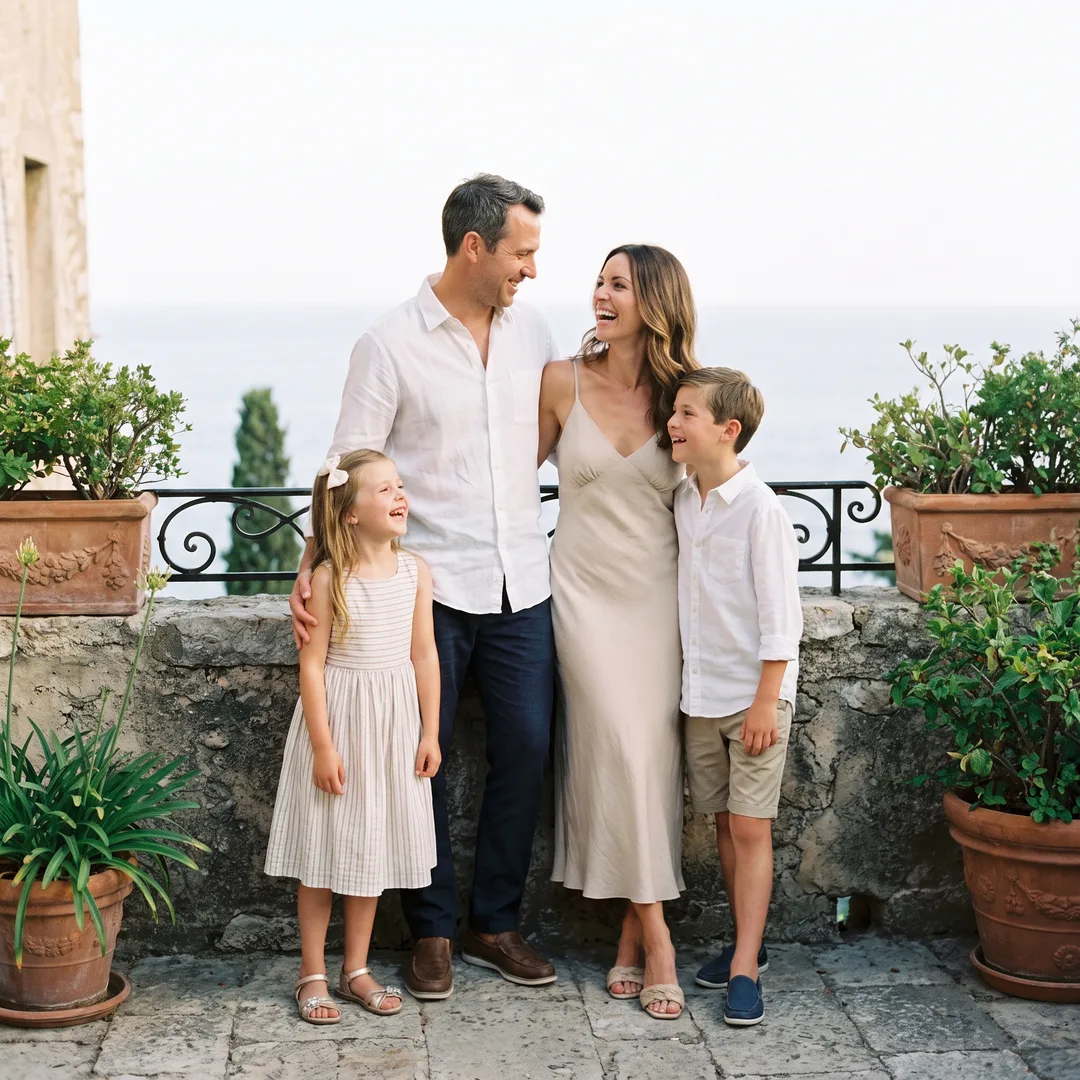 A family of four in smart casual spring clothes laughing together on a stone terrace above the Mediterranean in warm evening light
