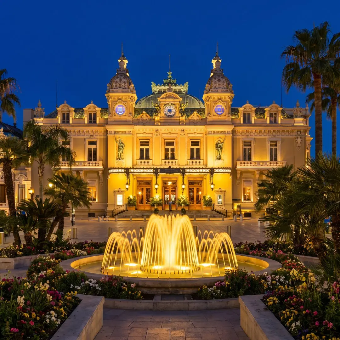 The Opéra de Monte-Carlo and Casino facade glowing gold at night, seen across the Place du Casino with palm trees, fountains and a dark blue sky