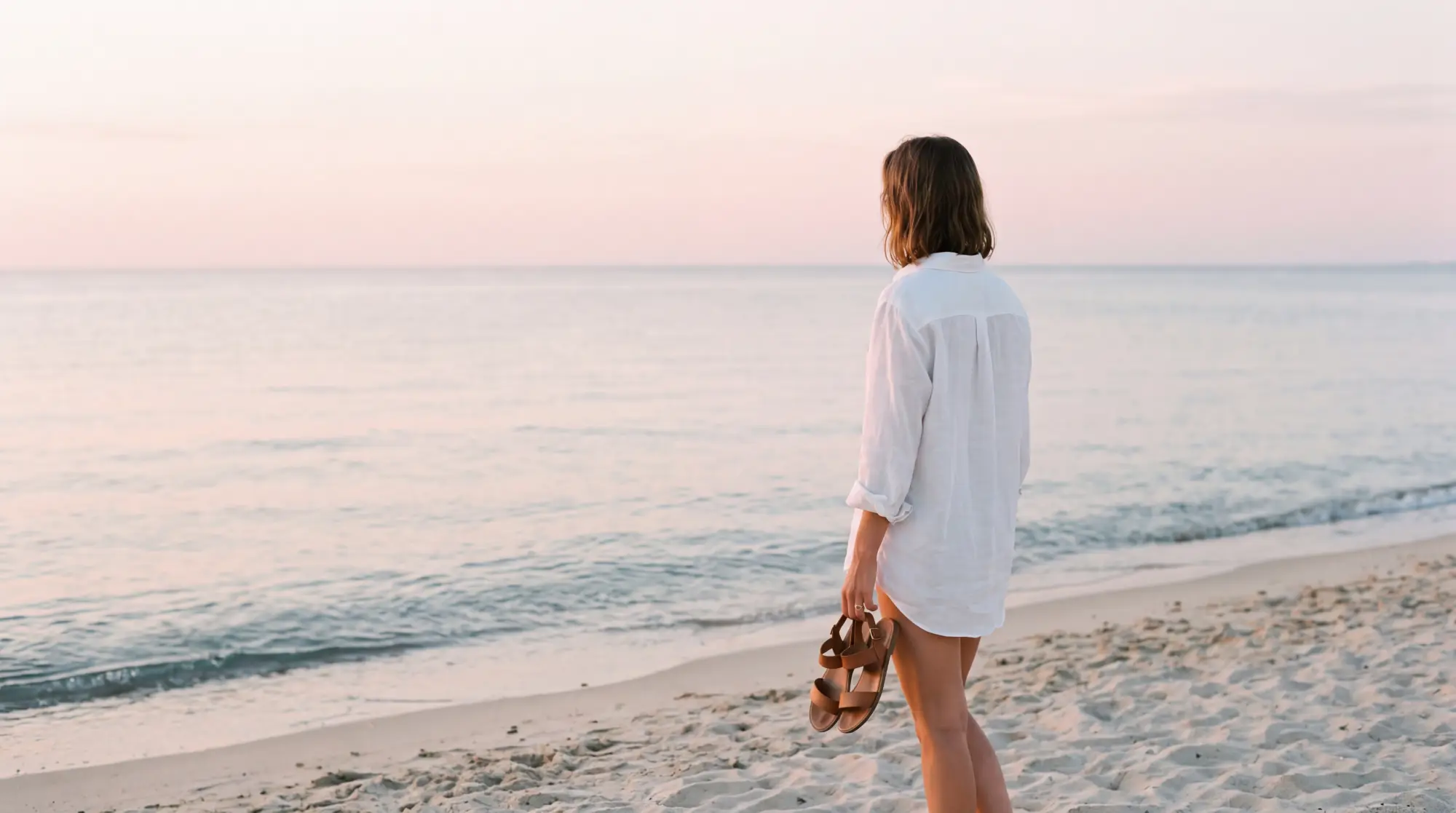 A woman in a white linen shirt walking alone at the water's edge on Pampelonne beach at sunrise