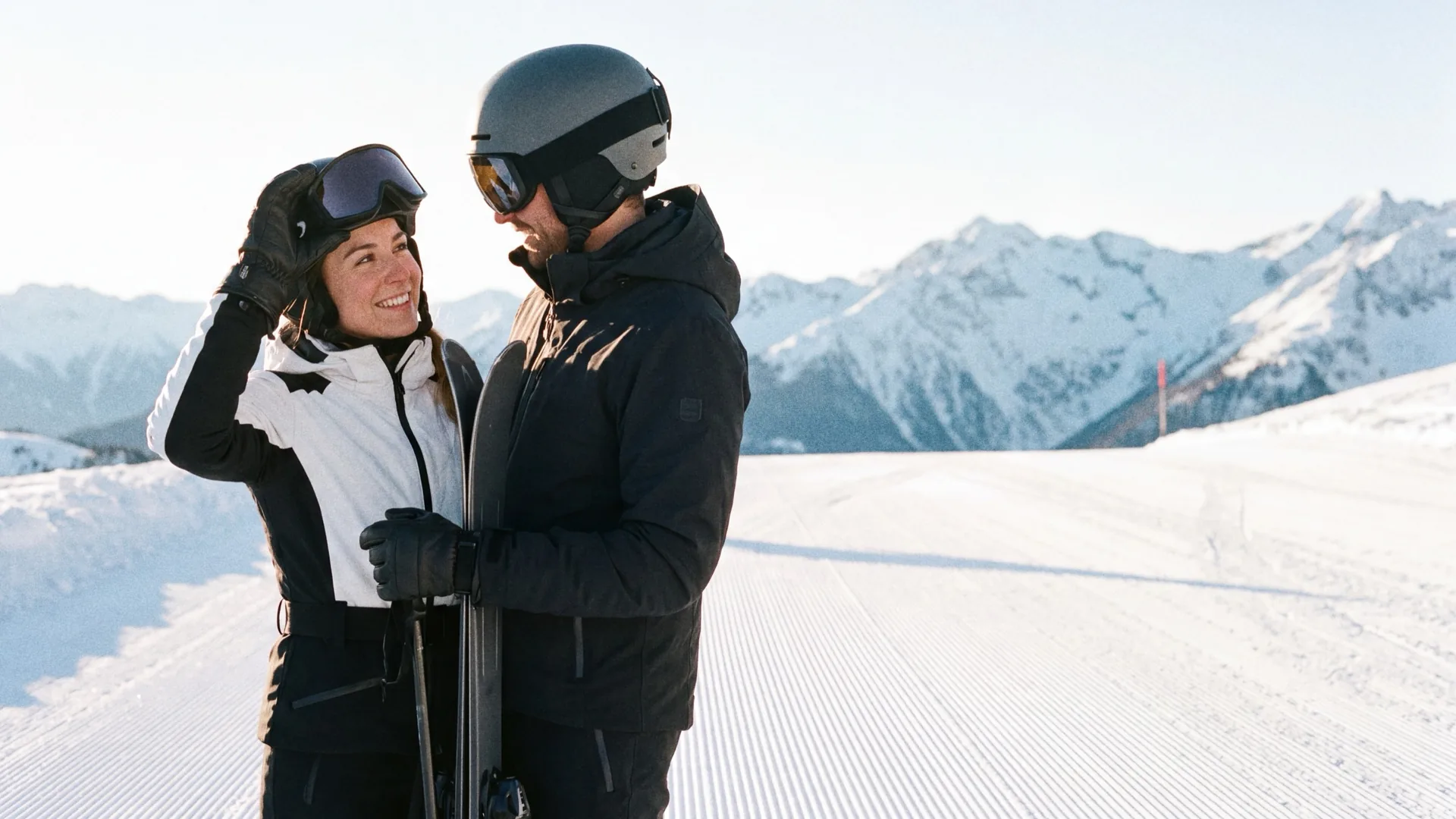 A couple in ski gear at the top of a groomed slope in late afternoon, she lifts her goggles and smiles at him, snowy peaks behind